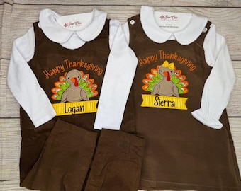 May include: Two matching Thanksgiving outfits. Each outfit includes a brown jumper with a white collared shirt and brown pants. The jumpers feature a turkey applique and the words "Happy Thanksgiving" and a name on a banner.