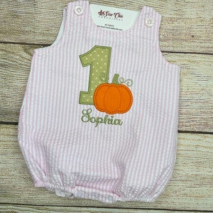 May include: Pink and white striped baby onesie with a green number one and a pumpkin applique. The onesie is embroidered with the name "Sophia".