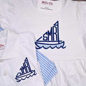 May include: White embroidered polo shirt and dress with blue sailboat design and the initials "SMR" on the sail. The dress has a blue and white striped trim.