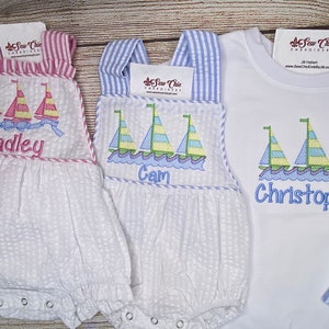 May include: White seersucker baby rompers and a white t-shirt with sailboat embroidery. The rompers have pink and blue striped accents and personalized names. The t-shirt has a blue name. The embroidery features sailboats with colorful sails.