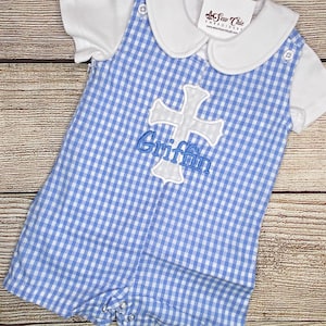 May include: A blue and white gingham baby romper with a white embroidered cross and the name "Griffin" on the front. The romper has a white Peter Pan collar and short sleeves.