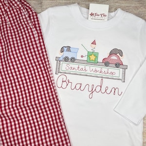 May include: A white long-sleeve shirt and red and white gingham pants. The shirt features an embroidered shelf with a blue train, a green jack-in-the-box, and a red car, with the text Santa's Workshop and the name Brayden embroidered below.