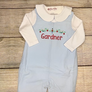 May include: A light blue baby romper with a white Peter Pan collar and long sleeves. The romper features an embroidered Christmas design with the name "Gardner" in red. The brand label "As Sew Chic" is visible.