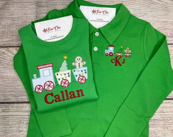 May include: Two green long-sleeved shirts. One is a crew neck with a train design and the name "Callan" embroidered in red. The other is a polo shirt with a train design and the initials "CKJ" embroidered in red. Both shirts have a Christmas theme.