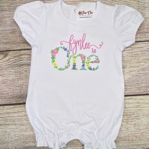 May include: White onesie with pink lettering that says "fynlee is one" with floral accents around the number one.