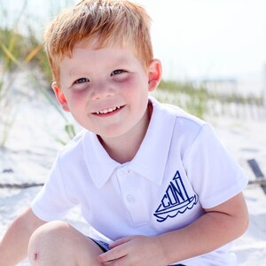 May include: A young boy with red hair smiles at the camera while wearing a white polo shirt with a blue sailboat design on the front. The boy is sitting on a sandy beach.