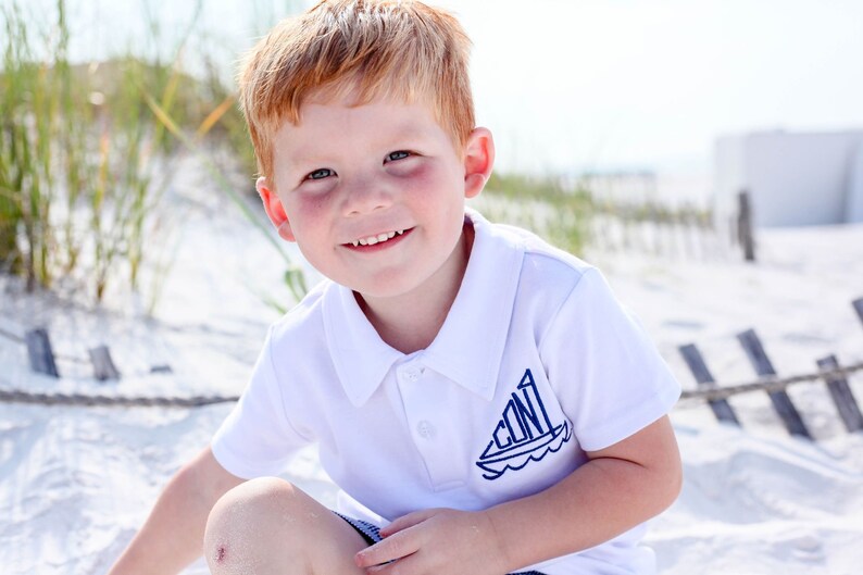 May include: A young boy with red hair smiles at the camera while wearing a white polo shirt with a blue sailboat design on the front. The boy is sitting on a sandy beach.