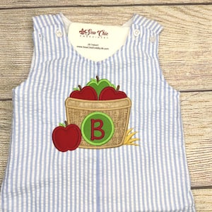 May include: Blue and white striped seersucker romper with an embroidered apple basket design. The basket contains red and green apples and a green letter "B". The romper has white buttons and the text "Sew Chic Embroidery".
