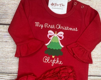 May include: A long-sleeved red shirt with ruffled cuffs and a ruffled hem. The front features an embroidered green Christmas tree topped with a pink bow, the text "My First Christmas" above, and the name "Blythe" embroidered below the tree.