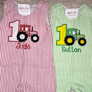 Peut inclure: Deux barboteuses en seersucker, une rouge et une verte, chacune avec une application de tracteur et le chiffre 1. La barboteuse rouge porte le nom "Jude", la verte "Sutton". Les deux ont des fermetures à boutons.