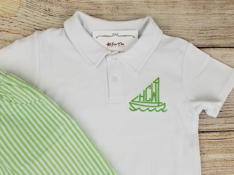 May include: White polo shirt with a green embroidered sailboat design featuring the initials "HCW".
