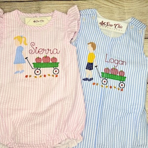 May include: Two seersucker rompers with embroidered designs. The pink romper features a girl, the name Sierra, and a wagon with apples. The blue striped romper features a boy, the name Logan, and a wagon with apples. Both have Sew Chie labels.