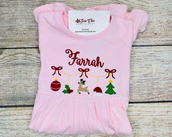May include: A light pink dress with short sleeves and a ruffled hem. The dress features an embroidered design with the name "Farrah" in burgundy script, along with Christmas ornaments and a reindeer.