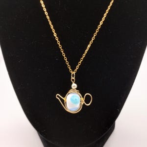 Könnte beinhalten: Eine goldfarbene Halskette mit einem Teekannen-Anhänger. Der Anhänger besteht aus goldenem Draht und enthält einen blau-weißen Stein. Die Halskette wird auf schwarzem Hintergrund präsentiert.