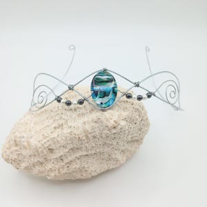 May include: A silver-toned wire crown with a central oval-shaped blue and green glass bead. The crown features a diamond-shaped design and is adorned with small black and white beads. The crown is displayed on a light-colored, textured surface.
