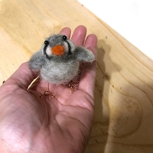 Puede incluir: Un pequeño pájaro de fieltro gris y blanco con un pico naranja y patas de alambre. El pájaro está sentado en la palma de una mano.