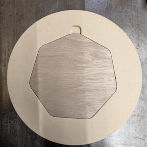 May include: A round, light beige wooden craft with a hexagonal cutout and a wooden insert. The insert has a light wood grain pattern. A small, curved tab is at the top of the craft.