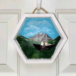 May include: A hexagonal white wooden frame containing a painted mountain landscape with a blue sky and birds. A dark brown nest with two small white birds with orange beaks sits in the foreground, surrounded by textured green moss.
