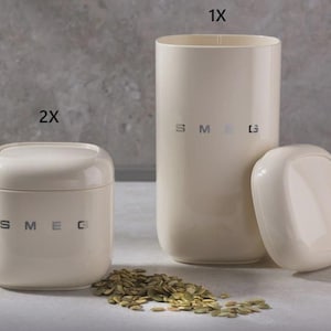 May include: Cream-colored kitchen canisters and utensil holder with the brand name "SMEG" in silver lettering. The set includes a small square canister with a lid, a tall cylindrical utensil holder, and a lid. A pile of seeds and a bundle of spaghetti are also visible.