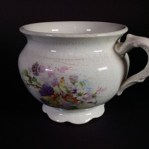 May include: Antique white ceramic bowl with a handle and floral design. The bowl features a scalloped base and a cracked glaze. The floral design includes purple berries, green leaves, and colorful flowers.