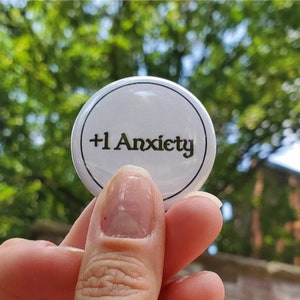 May include: White button with black text that reads " +1 Anxiety "