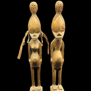 Op de afbeelding: Twee houten sculpturen van figuren met overdreven kenmerken, staande op houten bases. De figuren zijn gesneden in een gestileerde, tribale stijl en hebben grote hoofden met prominente ogen en monden. Beide figuren zijn zwanger en hebben lang, golvend haar.