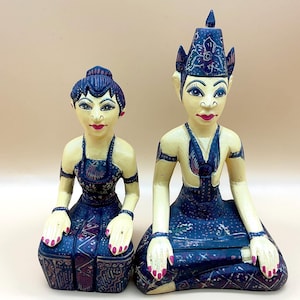 Può includere: Un paio di figurine in legno intagliate a mano che rappresentano un uomo e una donna in abiti tradizionali indonesiani. Le figurine sono dipinte in tonalità di blu, bianco e rosso, e presentano dettagli intricati. L'uomo indossa un cappello alto e appuntito, e la donna indossa un lungo abito fluido.