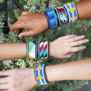 Pode incluir: Três pulseiras de contas com padrões geométricos coloridos. As pulseiras são feitas com contas azuis, amarelas, vermelhas, brancas e pretas. As pulseiras são usadas nos pulsos de três pessoas.