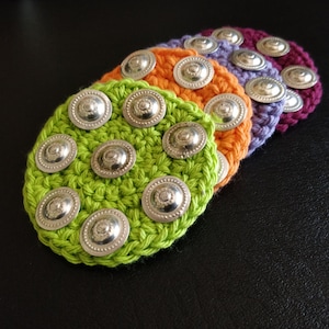 May include: Five crocheted coasters with silver metal buttons. The coasters are green, orange, purple, and two shades of orange.