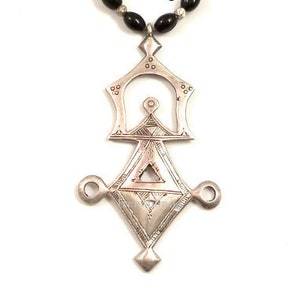 May include: A silver pendant necklace with a geometric design, featuring a central diamond shape with a smaller triangle inside. The pendant is attached to a black beaded chain with yellow and blue beads.