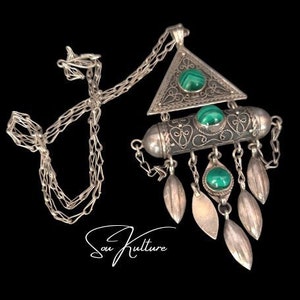 May include: A silver necklace with a triangular pendant featuring green stones and intricate filigree designs. The pendant is attached to a chain with a series of small, silver leaves dangling below.