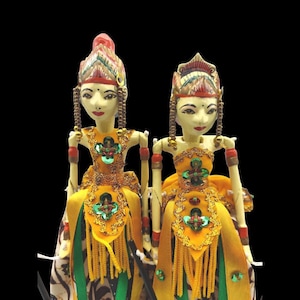 May include: Two traditional Indonesian puppets with intricate painted faces, elaborate headdresses, and colorful yellow and green costumes. The puppets are posed with their arms outstretched.