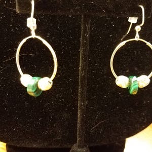 Op de afbeelding: Een paar zilveren oorringen. Elke oorring heeft een zilveren ring met drie kralen: twee crèmekleurige en één groene malachiet. De oorbellen worden gepresenteerd op een zwarte fluwelen achtergrond.