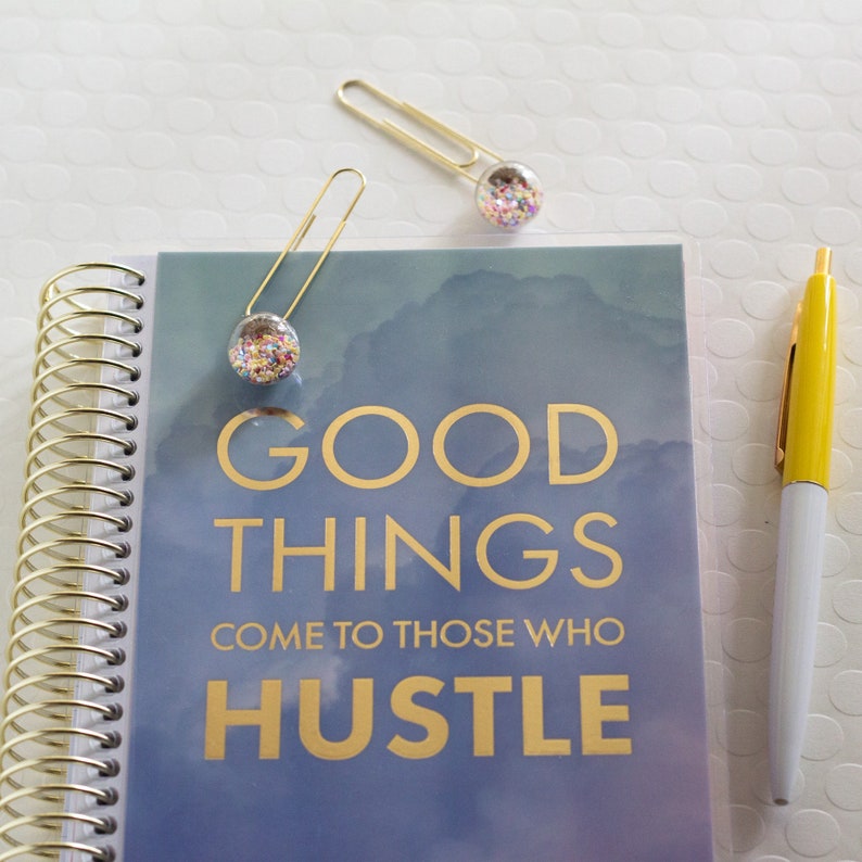 以下が含まれることがあります： 金色の螺旋状の綴じ込みと「GOOD THINGS COME TO THOSE WHO HUSTLE」という文字が書かれた青と白のノート。カラフルなコンフェッティで満たされた透明なガラスビーズが付いた金色のクリップが2つ、ノートに付けられています。
