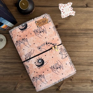 May include: A pink and peach fabric journal cover with a celestial design featuring hands, stars, moons, and a sun. The cover has a frayed edge and a brown leather tag that says "Lux Made".