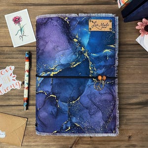 May include: A journal with a blue and purple marbled design, accented with gold flecks. It features a frayed edge, a black elastic closure with wooden beads and a peace sign charm. A small leather tag reads "Luv Made".