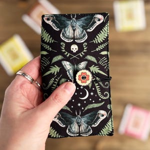 May include: A black fabric pouch with a zipper closure and a button closure. The pouch features a repeating pattern of moths, ferns, and a crescent moon on a black background.