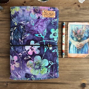 May include: A purple and blue fabric journal cover with a floral pattern. The cover has a brown leather tag with the text "Lux Made" on it. The journal is closed and has a black elastic band around it.