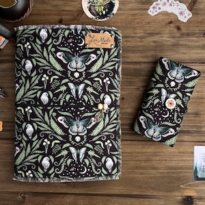 May include: A black and green fabric journal with a moth and fern pattern. The journal has a black elastic band and a small metal charm. The journal is closed and sitting on a wooden surface. The text "Live Made" is visible on the front cover.