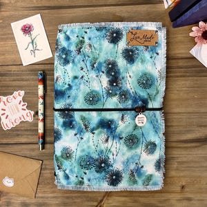 May include: A journal with a blue floral pattern, a black elastic band, and a wooden bead charm. A pen with a similar design, a sticker that says "Prove them wrong", and a small envelope are also visible.