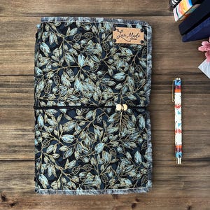 May include: A journal with a black and gold leaf pattern, secured with a black elastic band and wooden beads. A patterned pen and a small tag with the text "Luv Made" are also visible. The journal is on a wooden surface.