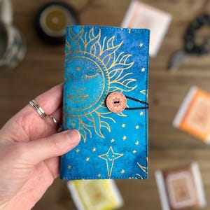 May include: A blue and gold celestial-themed journal cover with a sun and star design. The cover is secured with a button and elastic closure. The journal is held in a hand.