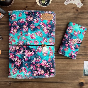 May include: Two turquoise blue fabric journals with a pink floral pattern. The larger journal has a black elastic band and a brown leather tag that says "Luv Made". The smaller journal has a brown button closure.