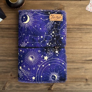 May include: A purple notebook with a celestial design featuring stars, constellations, and a crescent moon. The notebook has a black elastic closure and a brown leather tag that reads "Lux Made".