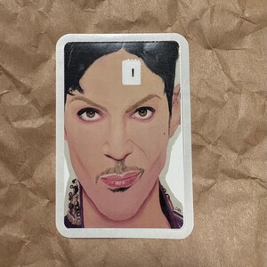 May include: A rectangular sticker featuring a portrait of a person with dark hair, a mustache, and a purple top. The portrait is a detailed illustration with a white border. The sticker has a small white rectangle on the forehead.