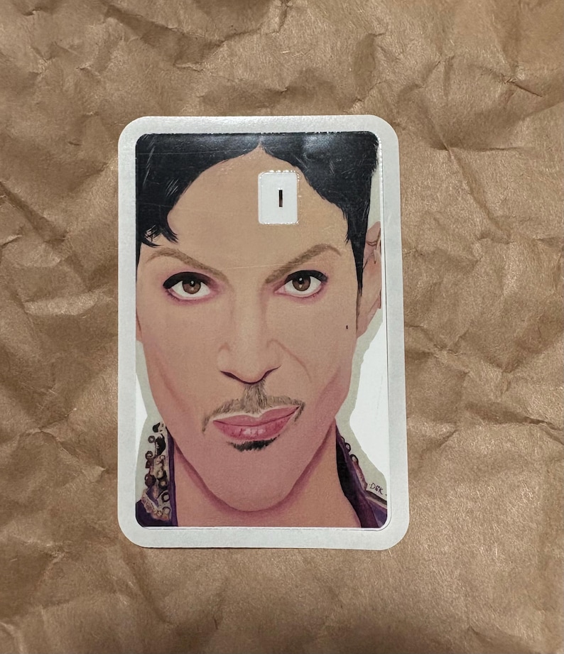 May include: A rectangular sticker featuring a portrait of a person with dark hair, a mustache, and a purple top. The portrait is a detailed illustration with a white border. The sticker has a small white rectangle on the forehead.