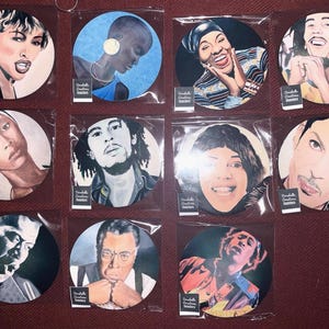 May include: A selection of round coasters, each with a portrait of a famous musician or performer. The coasters are individually packaged and feature various individuals, including Bob Marley and Prince. The text "Danielle Creations Inserts" is visible.