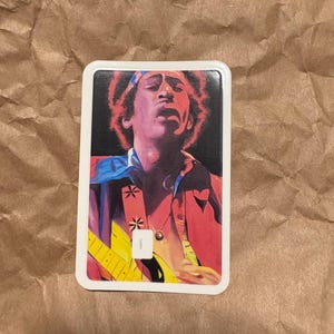 May include: Rectangular sticker featuring a vibrant illustration of a musician playing a yellow guitar. The image showcases the musician with a red jacket, blue accents, and a headband. The background is black.