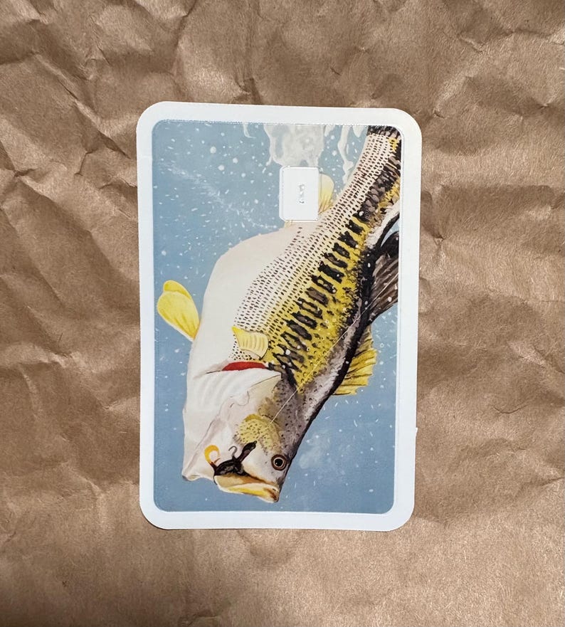 May include: A rectangular card featuring a detailed illustration of a fish, with yellow fins and a patterned body, set against a light blue background. The fish is positioned diagonally, with a hook near its mouth. The card has a white border.