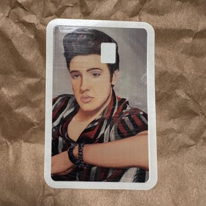 May include: A rectangular card featuring a portrait of a man with dark hair and a striped shirt in shades of red, black, and white. The man is wearing a black bracelet. The card has a white border.